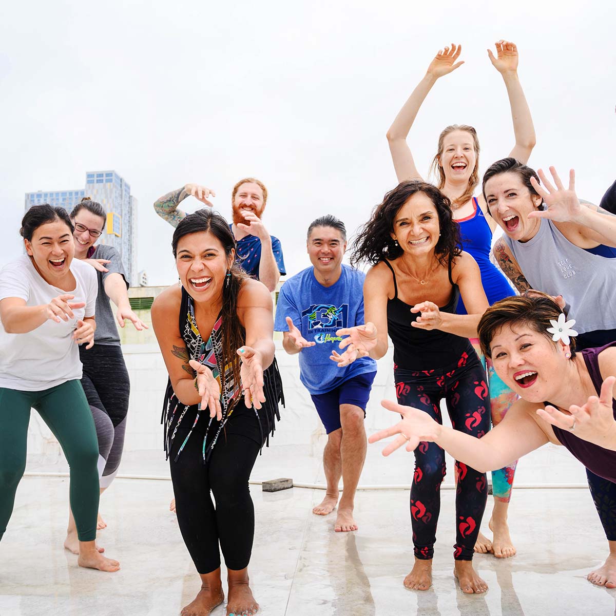 Mehrere Personen in sportlicher Kleidung stehen in einer dynamischen Pose mit ausgestreckten Armen und leicht gebeugten Knien auf einem hellen Boden vor einer Stadtkulisse und nehmen an einem Nia Kurs teil.